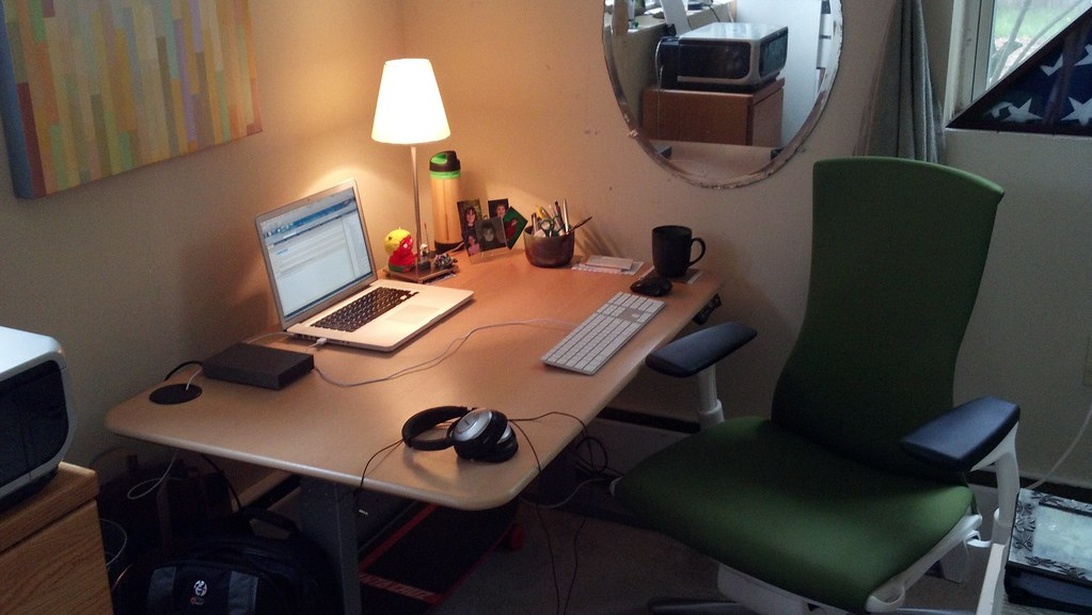 En mysig hemarbetsplats med träbord, laptop, externt tangentbord, hörlurar, skrivbordslampa och en bekväm grön ergonomisk stol. Personliga detaljer och omgivande belysning skapar en praktisk och personlig arbetsmiljö.