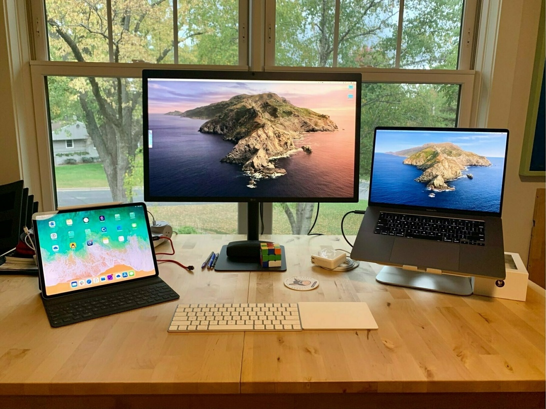 En ljus hemarbetsplats vid stora fönster med utsikt över träd. Arbetsplatsen har ett träbord med Apple-laptop, extern skärm, tangentbord och styrplatta, vilket främjar en produktiv och inbjudande arbetsmiljö tack vare det naturliga ljuset.